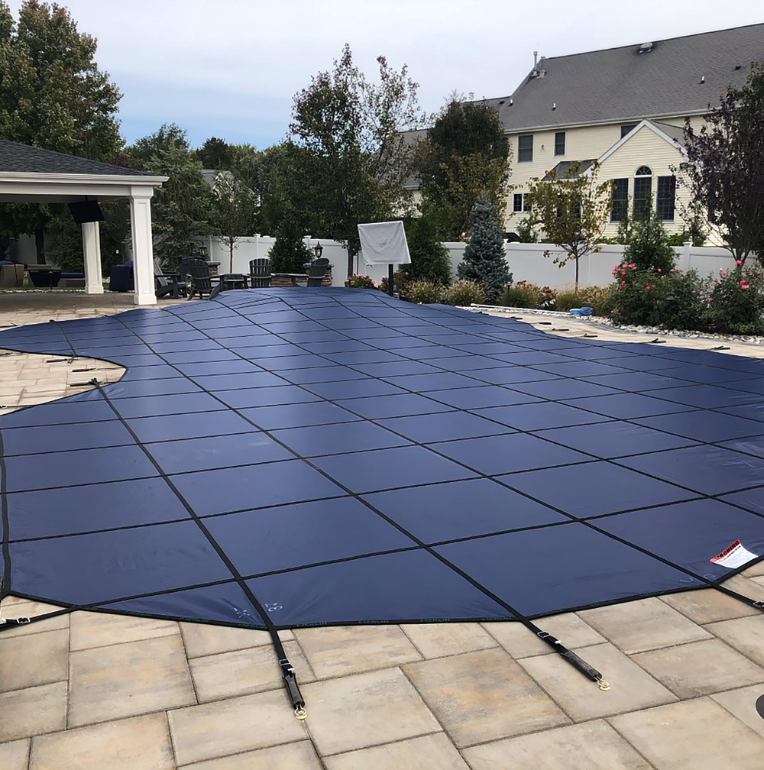 A backyard swimming pool covered with a blue safety cover, secured with black straps, surrounded by a stone patio and landscaped greenery, showcases Advanced Pool and Spa’s commitment to professional pool maintenance. A large house stands in the background.