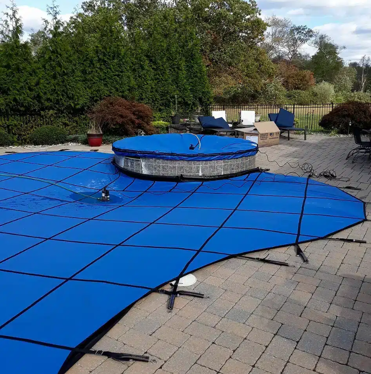 A backyard pool in St. Louis is covered with a bright blue safety cover from Advanced Pool and Spa, secured with black straps. A circular hot tub is also covered. Patio furniture and greenery complete the scene.